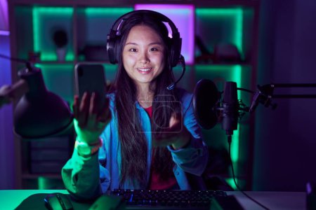 Foto de Mujer asiática joven jugando videojuegos con teléfono inteligente sonriendo amistoso ofreciendo apretón de manos como saludo y bienvenida. negocios exitosos. - Imagen libre de derechos