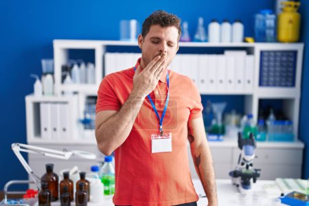 Foto de El joven hispano que trabajaba en el laboratorio científico se aburrió bostezando cansado cubriendo la boca con la mano. inquietud y somnolencia. - Imagen libre de derechos