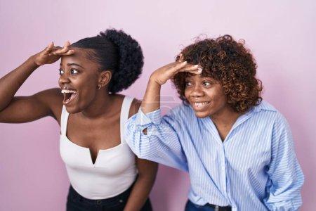 Foto de Dos mujeres africanas de pie sobre fondo rosa muy felices y sonrientes mirando lejos con la mano sobre la cabeza. concepto de búsqueda. - Imagen libre de derechos