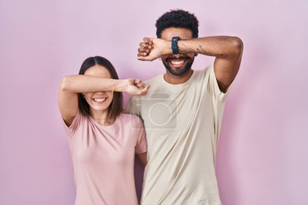 Foto de Joven pareja hispana juntos sobre fondo rosa sonriente alegre jugando a echar un vistazo a un boo con las manos mostrando la cara. sorprendido y salido - Imagen libre de derechos