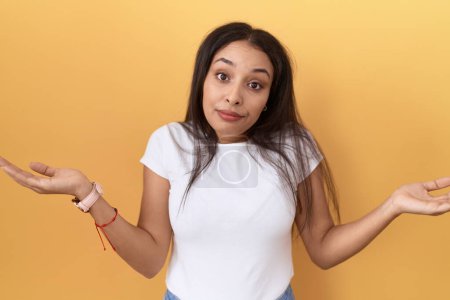 Photo for Young arab woman wearing casual white t shirt over yellow background clueless and confused expression with arms and hands raised. doubt concept. - Royalty Free Image