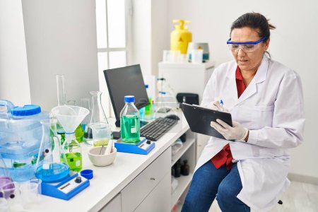 Foto de Mujer hispana de mediana edad vistiendo uniforme científico escribiendo en portapapeles en el laboratorio - Imagen libre de derechos