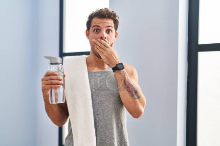 Photo for Young hispanic man wearing sportswear drinking water shocked covering mouth with hands for mistake. secret concept. - Royalty Free Image