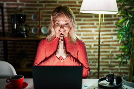 Téléchargez les photos : Femme blonde utilisant un ordinateur portable la nuit à la maison priant avec les mains ensemble demandant pardon souriant confiant. - en image libre de droit