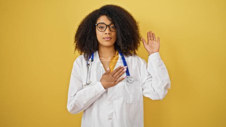 Foto de Médico afroamericano haciendo un juramento con la mano en el pecho sobre un fondo amarillo aislado - Imagen libre de derechos