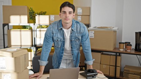 Téléchargez les photos : Jeune homme hispanique commerce électronique travailleur d'affaires en utilisant un ordinateur portable debout avec une expression sérieuse au bureau - en image libre de droit