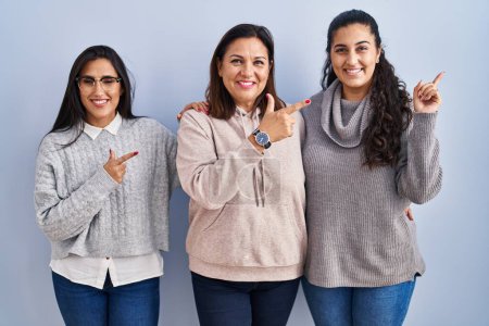 Photo for Mother and two daughters standing over blue background cheerful with a smile on face pointing with hand and finger up to the side with happy and natural expression - Royalty Free Image