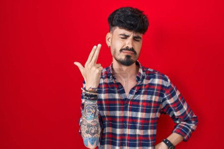 Foto de Joven hispano con barba de pie sobre fondo rojo disparando y matándose apuntando de la mano y los dedos a la cabeza como arma, gesto suicida. - Imagen libre de derechos