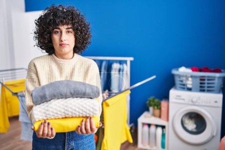 Photo for Young brunette woman with curly hair holding clean laundry relaxed with serious expression on face. simple and natural looking at the camera. - Royalty Free Image
