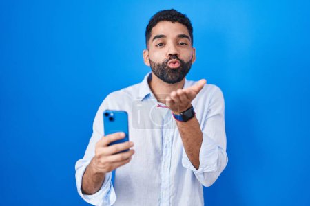 Foto de Hombre hispano con barba usando el mensaje de escritura de teléfonos inteligentes mirando a la cámara soplando un beso con la mano en el aire siendo encantador y sexy. expresión de amor. - Imagen libre de derechos