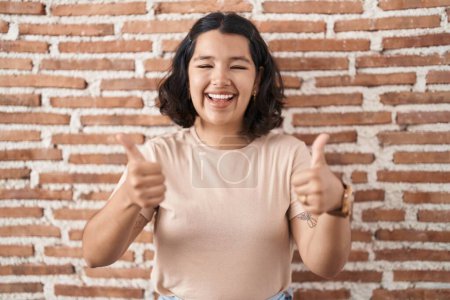 Foto de Mujer hispana joven de pie sobre ladrillos signo de éxito de la pared haciendo gesto positivo con la mano, pulgares hacia arriba sonriendo y feliz. expresión alegre y gesto ganador. - Imagen libre de derechos
