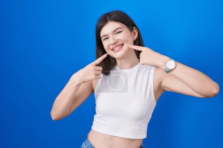 Téléchargez les photos : Young caucasian woman standing over blue background smiling cheerful showing and pointing with fingers teeth and mouth. dental health concept. - en image libre de droit