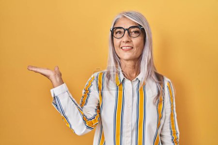 Photo for Middle age woman with grey hair standing over yellow background wearing glasses smiling cheerful presenting and pointing with palm of hand looking at the camera. - Royalty Free Image