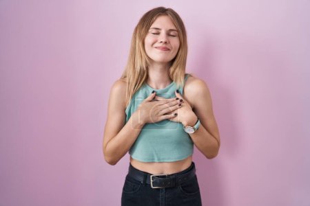 Téléchargez les photos : Blonde caucasian woman standing over pink background smiling with hands on chest with closed eyes and grateful gesture on face. health concept. - en image libre de droit