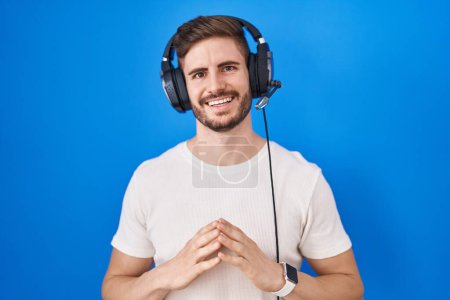 Foto de Hombre hispano con barba escuchando música usando auriculares con las manos juntas y los dedos cruzados sonriendo relajado y alegre. éxito y optimismo - Imagen libre de derechos