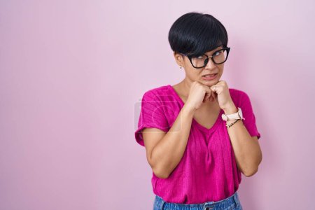 Foto de Joven mujer asiática con el pelo corto de pie sobre el fondo rosa riendo nervioso y emocionado con las manos en la barbilla mirando a un lado - Imagen libre de derechos