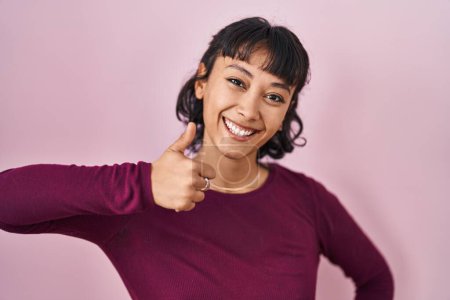 Foto de Young beautiful woman standing over pink background doing happy thumbs up gesture with hand. approving expression looking at the camera showing success. - Imagen libre de derechos