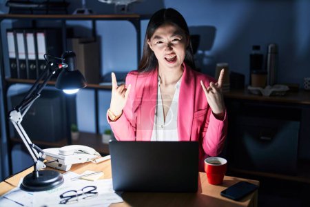 Foto de Mujer joven china que trabaja en la oficina por la noche gritando con expresión loca haciendo símbolo de roca con las manos arriba. Estrella de la música. concepto pesado. - Imagen libre de derechos