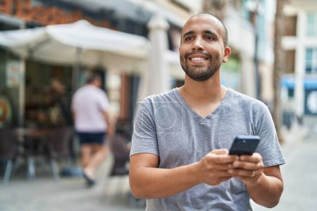 Téléchargez les photos : Jeune homme latino souriant confiant en utilisant smartphone à la terrasse du café - en image libre de droit