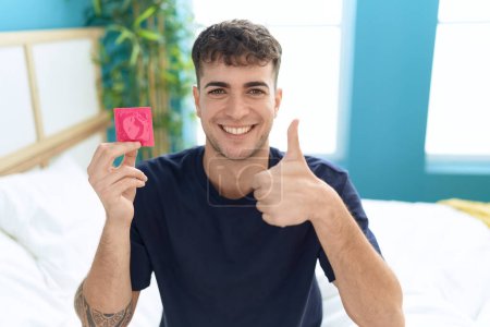 Téléchargez les photos : Jeune homme hispanique assis sur le lit tenant préservatif souriant heureux et positif, pouce levé faire excellent et signe d'approbation - en image libre de droit