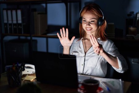 Foto de Joven mujer caucásica trabajando en la oficina por la noche mostrando y señalando hacia arriba con los dedos número ocho mientras sonríe confiado y feliz. - Imagen libre de derechos