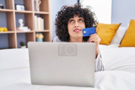Téléchargez les photos : Jeune femme du Moyen-Orient utilisant ordinateur portable et carte de crédit couché sur le lit dans la chambre - en image libre de droit