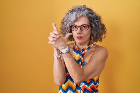 Téléchargez les photos : Femme d'âge moyen avec les cheveux gris debout sur fond jaune tenant pistolet symbolique avec geste de la main, jouant tuer armes de tir, visage en colère - en image libre de droit