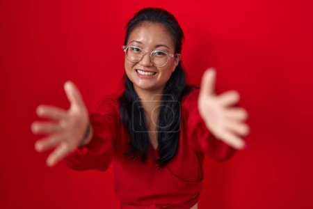 Téléchargez les photos : Asiatique jeune femme debout sur fond rouge regardant la caméra souriant avec les bras ouverts pour étreindre. expression joyeuse embrassant le bonheur. - en image libre de droit