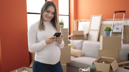 Photo for Young pregnant woman using smartphone standing at new home - Royalty Free Image