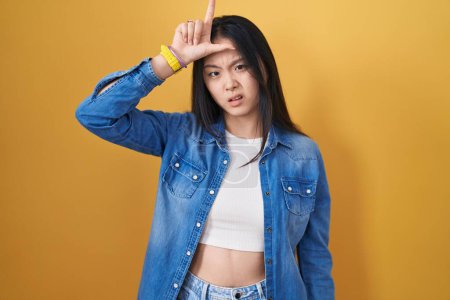 Photo for Young asian woman standing over yellow background making fun of people with fingers on forehead doing loser gesture mocking and insulting. - Royalty Free Image