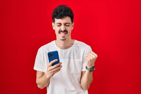 Photo for Hispanic man using smartphone over red background very happy and excited doing winner gesture with arms raised, smiling and screaming for success. celebration concept. - Royalty Free Image