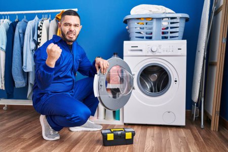 Photo for Hispanic repairman working on washing machine annoyed and frustrated shouting with anger, yelling crazy with anger and hand raised - Royalty Free Image