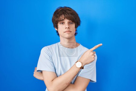 Téléchargez les photos : Hispanic young man standing over blue background pointing with hand finger to the side showing advertisement, serious and calm face - en image libre de droit