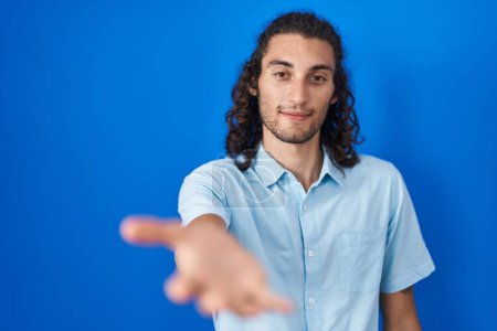 Photo for Young hispanic man standing over blue background smiling cheerful offering palm hand giving assistance and acceptance. - Royalty Free Image