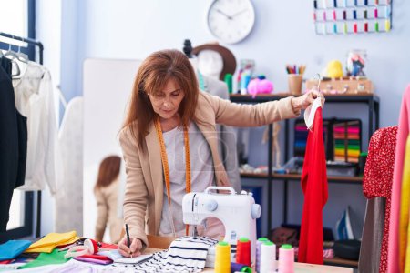 Téléchargez les photos : Middle age woman tailor holding t shirt writing on notebook at clothing factory - en image libre de droit