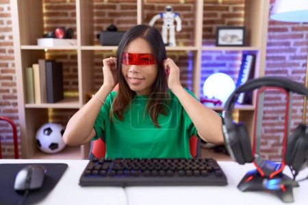 Foto de Mujer china de mediana edad con gafas de realidad virtual que cubren las orejas con los dedos con expresión molesta por el ruido de la música alta. concepto de sordo. - Imagen libre de derechos