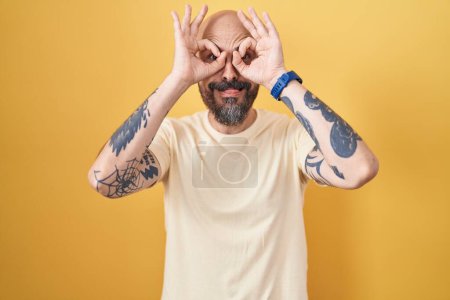 Téléchargez les photos : Homme hispanique avec des tatouages debout sur fond jaune faisant un geste correct comme des jumelles sortant la langue, les yeux regardant à travers les doigts. expression folle. - en image libre de droit