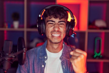 Foto de Joven hispano jugando videojuegos celebrando sorprendido y sorprendido por el éxito con los brazos levantados y los ojos cerrados - Imagen libre de derechos