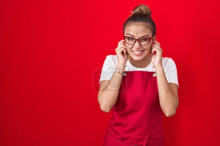 Foto de Mujer hispana joven con delantal de camarera sobre fondo rojo cubriendo las orejas con los dedos con expresión molesta por el ruido de la música alta. concepto de sordo. - Imagen libre de derechos