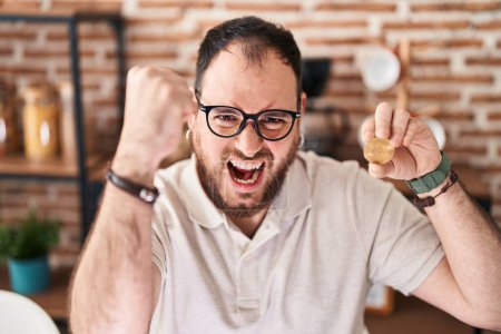 Téléchargez les photos : Plus taille homme hispanique avec barbe tenant monnaie virtuelle Bitcoin ennuyé et frustré criant avec colère, hurlant fou de colère et la main levée - en image libre de droit