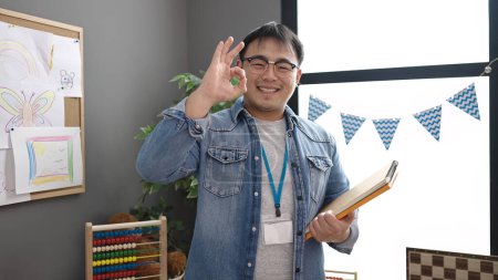 Foto de Joven profesor chino sosteniendo libros haciendo buen gesto en el jardín de infantes - Imagen libre de derechos
