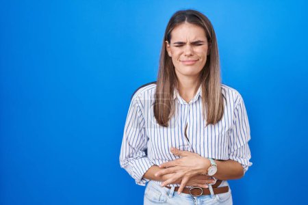 Photo for Hispanic young woman standing over blue background with hand on stomach because indigestion, painful illness feeling unwell. ache concept. - Royalty Free Image
