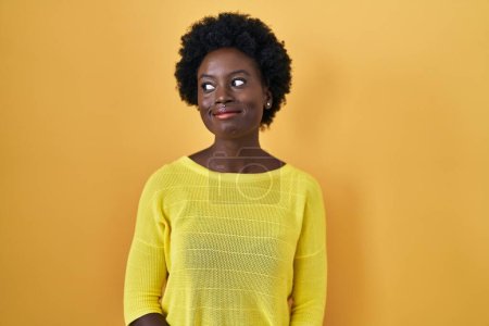 Foto de Mujer joven africana de pie sobre el estudio amarillo sonriendo mirando hacia un lado y mirando hacia otro pensando. - Imagen libre de derechos