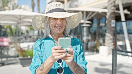 Foto de Turista mujer de pelo gris de mediana edad sonriendo confiada usando teléfono inteligente en la terraza de la cafetería - Imagen libre de derechos