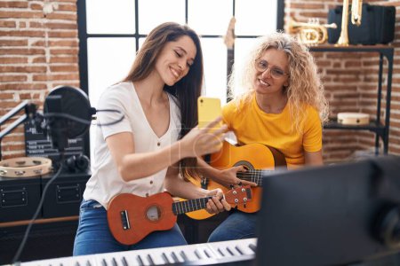 Téléchargez les photos : Deux femmes musiciens jouant de la guitare classique et de l'ukulélé regardant l'écran du smartphone au studio de musique - en image libre de droit