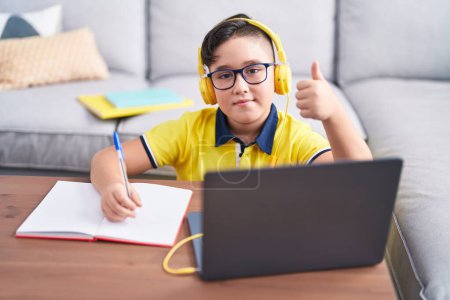 Téléchargez les photos : Jeune hispanique enfant faire ses devoirs avec ordinateur portable à la maison souriant heureux et positif, pouce levé faire excellent et signe d'approbation - en image libre de droit