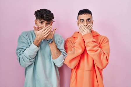 Foto de Joven pareja gay hispana de pie sobre fondo rosa impactó cubriendo la boca con las manos por error. concepto secreto. - Imagen libre de derechos