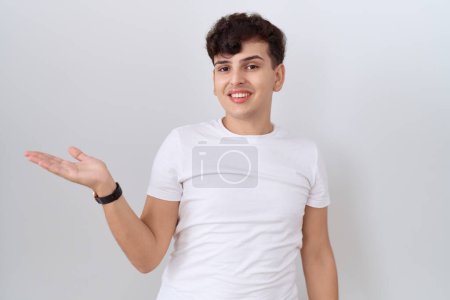 Foto de Joven hombre no binario vistiendo casual camiseta blanca sonriendo alegre presentando y señalando con la palma de la mano mirando a la cámara. - Imagen libre de derechos