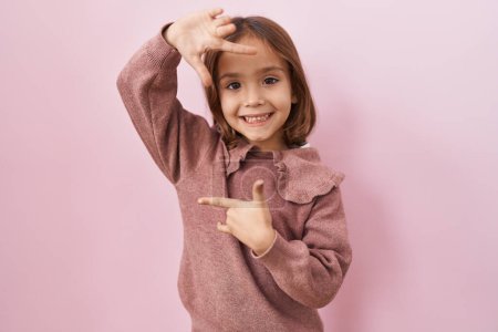 Téléchargez les photos : Petite fille hispanique debout sur fond rose souriant faisant cadre avec les mains et les doigts avec le visage heureux. concept de créativité et photographie. - en image libre de droit