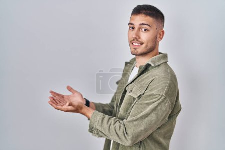 Téléchargez les photos : Jeune homme hispanique debout sur fond isolé pointant de côté avec les mains ouvertes paumes montrant l'espace de copie, présentant la publicité souriant heureux excité - en image libre de droit